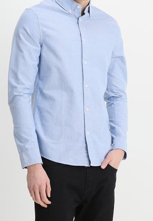 Mężczyzna w jasnoniebieskiej koszuli na guziki z kołnierzykiem i czarnych spodniach, stoi z jedną ręką w kieszeni na jednolitym tle.