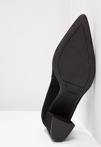 Scarpa nera in suede con punta a punta e tacco alto in gomma testurizzata, mostrata dal lato della suola su uno sfondo angolare bianco.