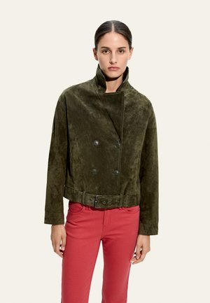 Kobieta o ciemnych włosach w zielonej zamszowej marynarce dwurzędowej z paskiem i czerwonych spodniach, stojąca na jasnobeżowym tle.