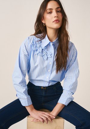 Frau mit langen braunen Haaren, die ein hellblau gestreiftes Hemd mit Blumenapplikationen und dunkelblaue Jeans trägt, sitzt auf einer Holzkiste mit verschränkten Händen.