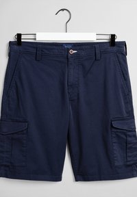 Short cargo bleu marine avec poches à rabat latérales, passants de ceinture, fermeture par bouton, suspendu sur un cintre blanc et noir sur un fond uni.