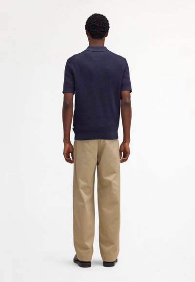 Mies seisoo selin päin, yllään laivastonsininen lyhythihainen poolopaita, beiget leveälahkeiset housut ja mustat kengät, yksinkertaista taustaa vasten.