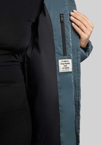 Giacca imbottita blu e nera con una tasca interna con zip e un'etichetta che recita "Un abbraccio caldo fatto di tessuto." Esterno liscio e lucido.