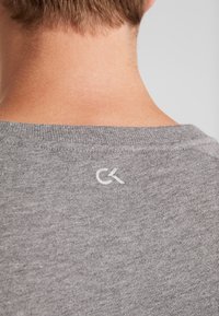 Vue de dos d'une personne portant un t-shirt gris avec un petit logo blanc "CK" au centre supérieur du dos, près de l'encolure.