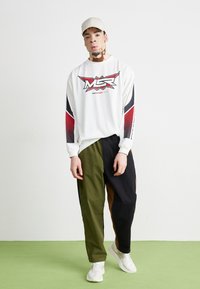 Weißer Sweater mit rographic und schwarzen Akzenten, kombiniert mit color-blocked Hosen in Oliv und Schwarz. Weiße Sneakers und beigefarbene Mütze.