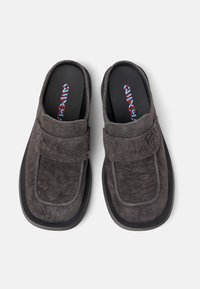 Zapatos slip-on grises y texturizados con una suela gruesa negra y una parte superior suave que presenta una correa de velcro. El interior muestra plantillas de marca en azul y rojo.