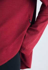 Bordo ilgi piedurkņu tops ar ribotu tekstūru, ar sānu kabatu un diskretām šuvēm, parādīts uz gaiša fona.