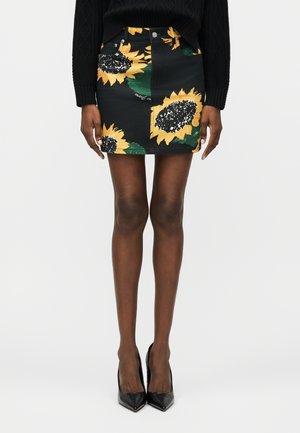 Oseba, oblečena v črno mini krilo z velikimi vzorci sončnic, črn pulover in črne koničaste visoke pete, stoji na enobarvni podlagi.