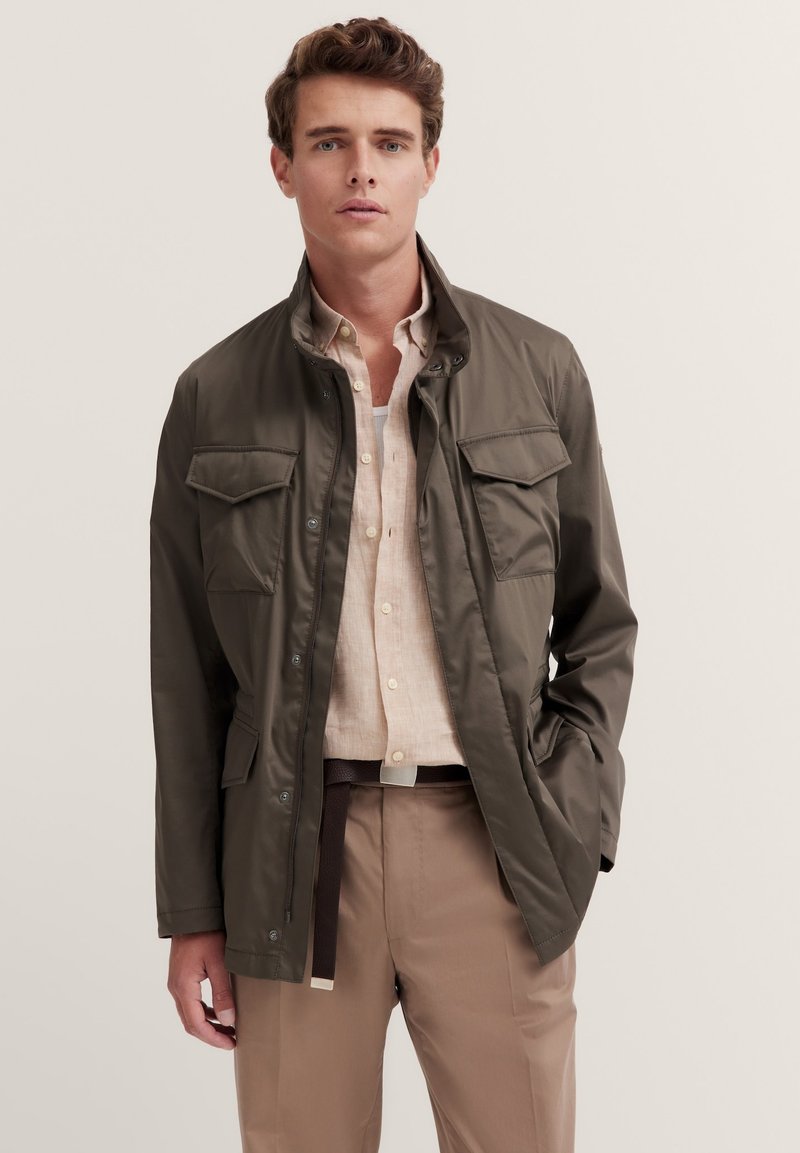Olivgrüne Jacke mit einem eleganten Design, zwei Vordertaschen und einem Knopfverschluss. Getragen über einem hellen Hemd und beigen Hosen.