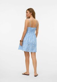 Robe fleurie bleue avec des fines bretelles, dos smocké et jupe évasée. Présente une texture légère et un motif de feuilles blanches. Associée à des sandales noires.