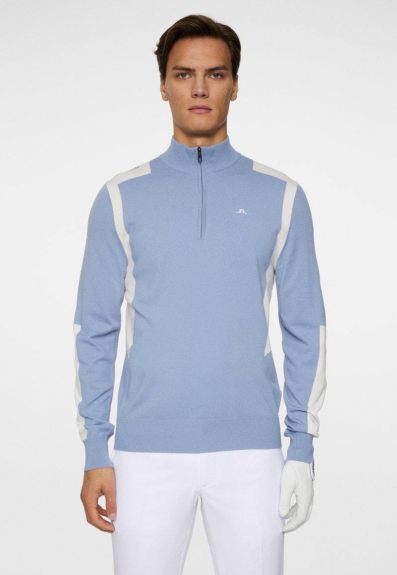 Ljusblå zip-tröja med vita axelpaneler. Mjuk textur, figurnära design, ribbade manschetter och en logotyp på bröstet. Vita handskar på.