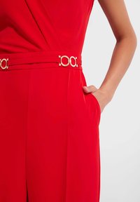 Czerwony kombinezon z zapięciem w talii, gładkiego materiału, szerokim paskiem z złotym okrągłym elementem oraz kieszeniami po bokach.