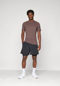 En atletisk mann står mot en enkel bakgrunn iført en brun kortermet skjorte, svarte lagdelte shorts, hvite sokker og hvite joggesko.