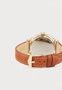 Bracelet en cuir brun avec un boîtier et une boucle en acier inoxydable doré. Présente des coutures visibles et un verre rond, affichant "TOUT EN ACIER INOXYDABLE."