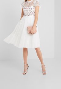 Valkoinen mekko, jossa on kukkaistikkauksia, läpikuultavia pitsiyksityiskohtia ja tyllihame. Yhdistetty hopeisiin remmikorkokenkiin ja pieneen vaaleanpunaiseen iltalaukkuun.