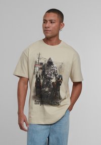 Beige bavlněné tričko s grafikou postavy v kapuci se dvěma psy a textem "zapomenuté tváře" v černé barvě na spodní části.