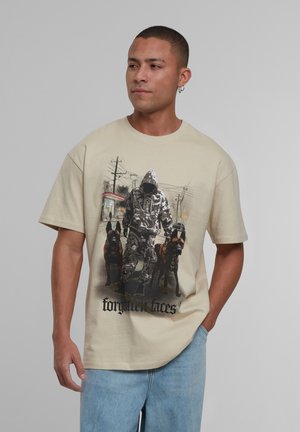 Beige kokvilnas t-krekls ar grafiku, kurā attēlots cilvēks kapucē ar diviem suņiem, un apakšā melnā krāsā ir teksts "aizmirstas sejas".