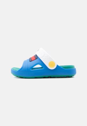 Blå og grønn sandsandal med en hvit rem som har en gul rund knapp og røde og svarte dekorative elementer.