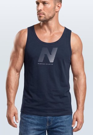 Muskelbepackter Mann trägt ein marineblaues Tanktop mit einem gepunkteten „N“ und dem Schriftzug „Everyday Athletics“, dazu blaue Jeans, blickt zur Seite.