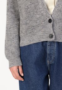 Cardigan grigio a coste realizzato in materiale morbido, dotato di scollatura a V, due bottoni neri e maniche ampie, abbinato a jeans denim blu scuro.