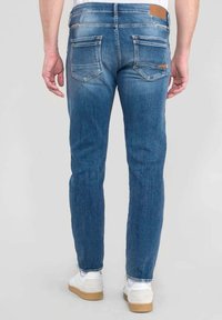 Mies, który ma na sobie dopasowane niebieskie dżinsy i białe sneakersy, stoi plecami do ujęcia na jednolitym jasnoszarym tle.