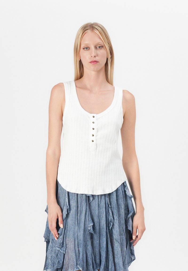 Hvit ribbet tanktop med rund hals, har fem knapper foran og en lett bølget kant. Kombinert med et blått volangskjørt.