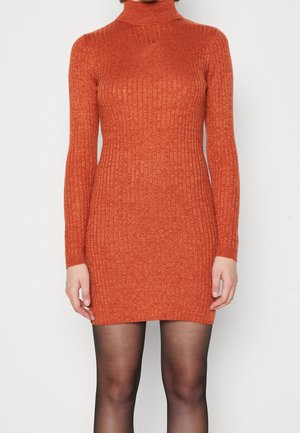 Femme portant une robe orange moulante à col roulé côtelée à manches longues avec des collants noirs transparents, debout devant un fond blanc uni.