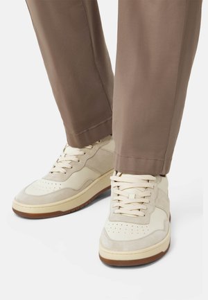 Sneakers bianchi con pannelli in suede e pelle, dotati di una punta perforata e lacci beige, abbinati a pantaloni marroni, leggermente svasati.