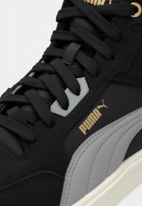 Baskets Puma noires et grises avec lacets noirs, logo Puma doré et semelle blanche visibles en gros plan.