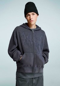 Mladostnik nosi črn beanie in prenaglašeno temno sivo zadrgo s kapuco s teksturirano grafiko ter ima roke v žepih.