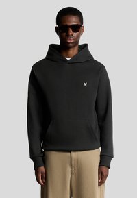 Sort hættetrøje med en frontlomme og snøre. Har et sølvfarvet logo på brystet. Bæres med beige teksturerede bukser.