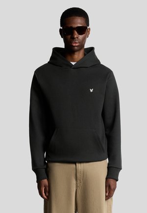 Svart hoodie med framficka och snörning i huvan. Har en silverlogotyp på bröstet. Bärs med beiga texturerade byxor.