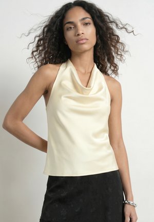 Vrouw met krullend haar, die een mouwloze crèmekleurige satijnen top en een zwarte rok draagt, staat met één gebogen arm en draagt een chunky armband.