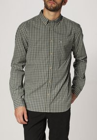 Mężczyzna w zielonej i szarej koszuli w kratę z długimi rękawami oraz czarnych spodniach, stojący z jedną ręką w kieszeni.