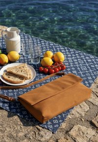 Μπλε υφασμάτινο σχέδιο πάνω σε πέτρα δίπλα σε καθαρό νερό με καφέ δερμάτινη τσάντα, λεμόνια, ντοματίνια, ψωμί σε πιάτο, λευκό κανάτα και δύο άδεια ποτήρια.