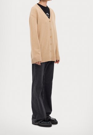 Modèle portant un cardigan beige boutonné sur le devant par-dessus un T-shirt noir, un jean noir ample et des mocassins imposants.