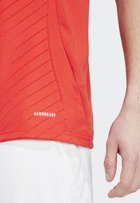 Červené športové tričko vyrobené z priedušnej tkaniny s diagonálnym pruhom. Logo "AEROREADY" potlačené na bielom pozadí pri okraji. Biele šortky sú čiastočne viditeľné.