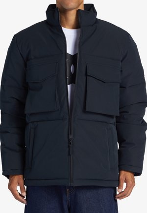 Navy modrá puffer bunda s vysokým límcem, zipem vpředu a čtyřmi velkými předními kapsami. Vyrobena z hladkého, izolačního materiálu.