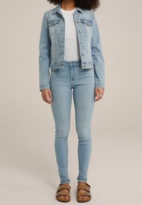 Ljusblå denimjacka med två bröstfickor, vit topp och ljusblå smalbyxor. Bruna sandaler med remmar. Slät tygtextur.