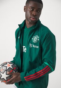 Chaqueta verde con mangas a rayas rojas, logo del Manchester United y texto blanco. Sosteniendo un balón de fútbol con un patrón en blanco y negro.