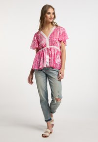 Blusa rosa a fantasia con maniche a cappuccio e scollatura a V, cintrata in vita con un fiocco. Abbinata a jeans azzurri strappati e sandali.