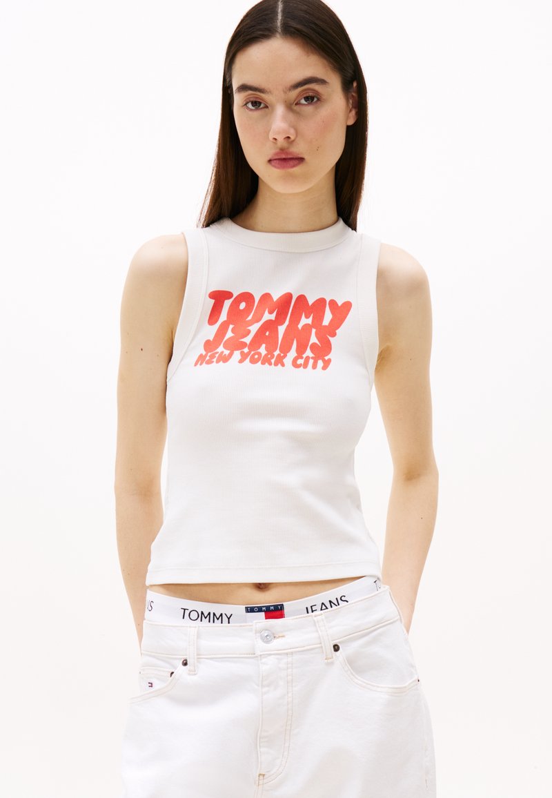 Valge ilma varrukateta lühike topp, millel on punane "TOMMY JEANS" tekst ja "NEW YORK CITY" trükk. Kombineeritud valgete teksadega, millel on brändi logo taljeosa.