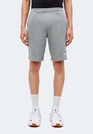 Bărbat purtând pantaloni scurți gri Puma, adidași albi Puma și șosete albe până la jumătatea gambei, stând cu brațele relaxate pe lângă corp, pe un fundal simplu.