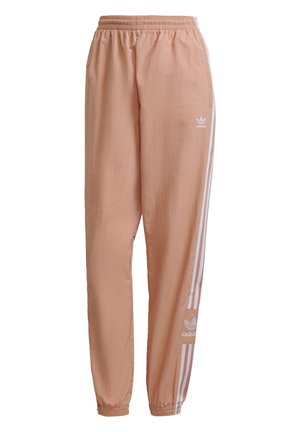 Osoba w brzoskwiniowych spodniach dresowych Adidas z białymi bocznymi paskami i białych trampkach, stojąca na tle jednolitego jasnego tła.