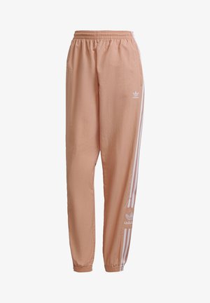 Osoba w brzoskwiniowych spodniach dresowych Adidas z białymi bocznymi paskami i białych trampkach, stojąca na tle jednolitego jasnego tła.