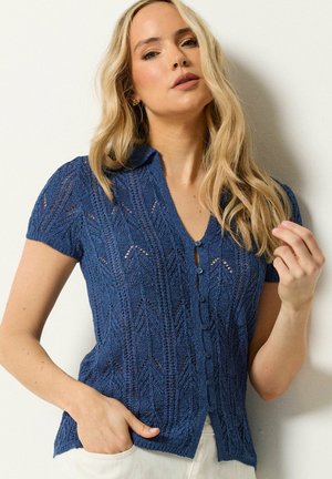 Kobieta o długich blond włosach, ubrana w niebieską, krótko rękawową dzianinową bluzkę na guziki oraz jasne spodnie, pozująca z jedną ręką w kieszeni.