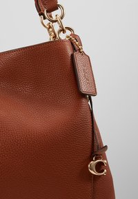 Sac à main en cuir marron texturé avec des ferrures dorées, une étiquette attachée et un charm doré en forme de "C" sur la sangle latérale.