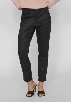 Pantalones negros ajustados con textura suave, bolsillos delanteros y puños enrollados. Combinados con zapatos de tacón alto negros de punta abierta.
