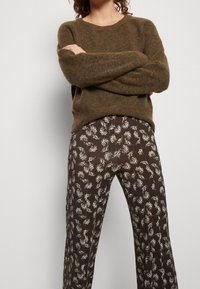 Osoba v hnědém chlupatém svetrě a vysoce pasovaných hnědých kalhotách s bílým vzorem připomínajícím listy, se zkříženýma rukama na hrudi.