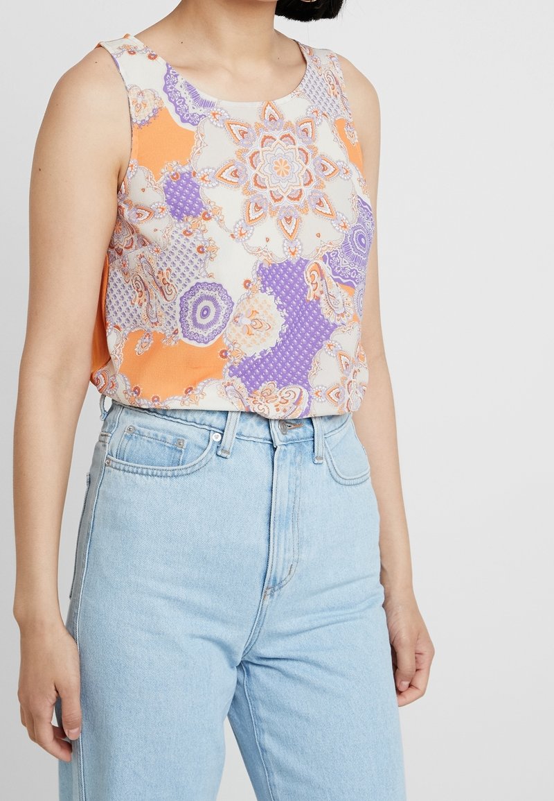 Top sin mangas de tejido ligero con un patrón geométrico en colores naranja, púrpura y crema. Combinado con unos vaqueros de tiro alto en azul claro.